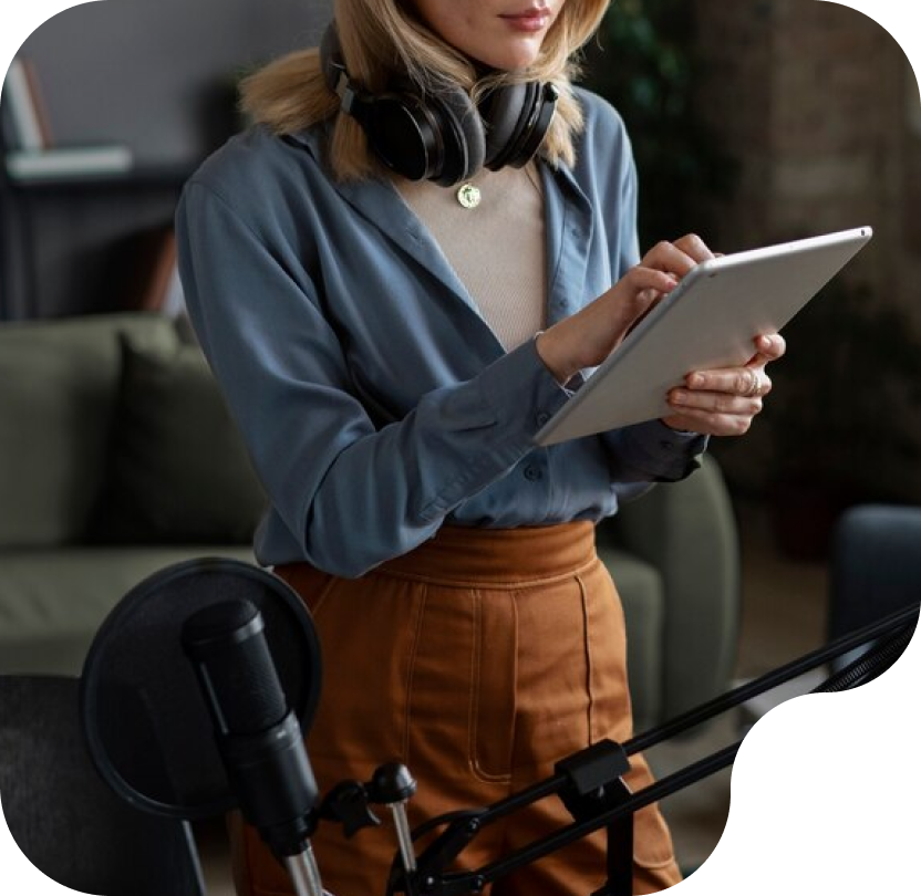 Woman wearing headphones using a tablet in a podcast recording setup, featuring a microphone and a comfortable studio environment, reflecting PodcastCrew's professional audio production services.