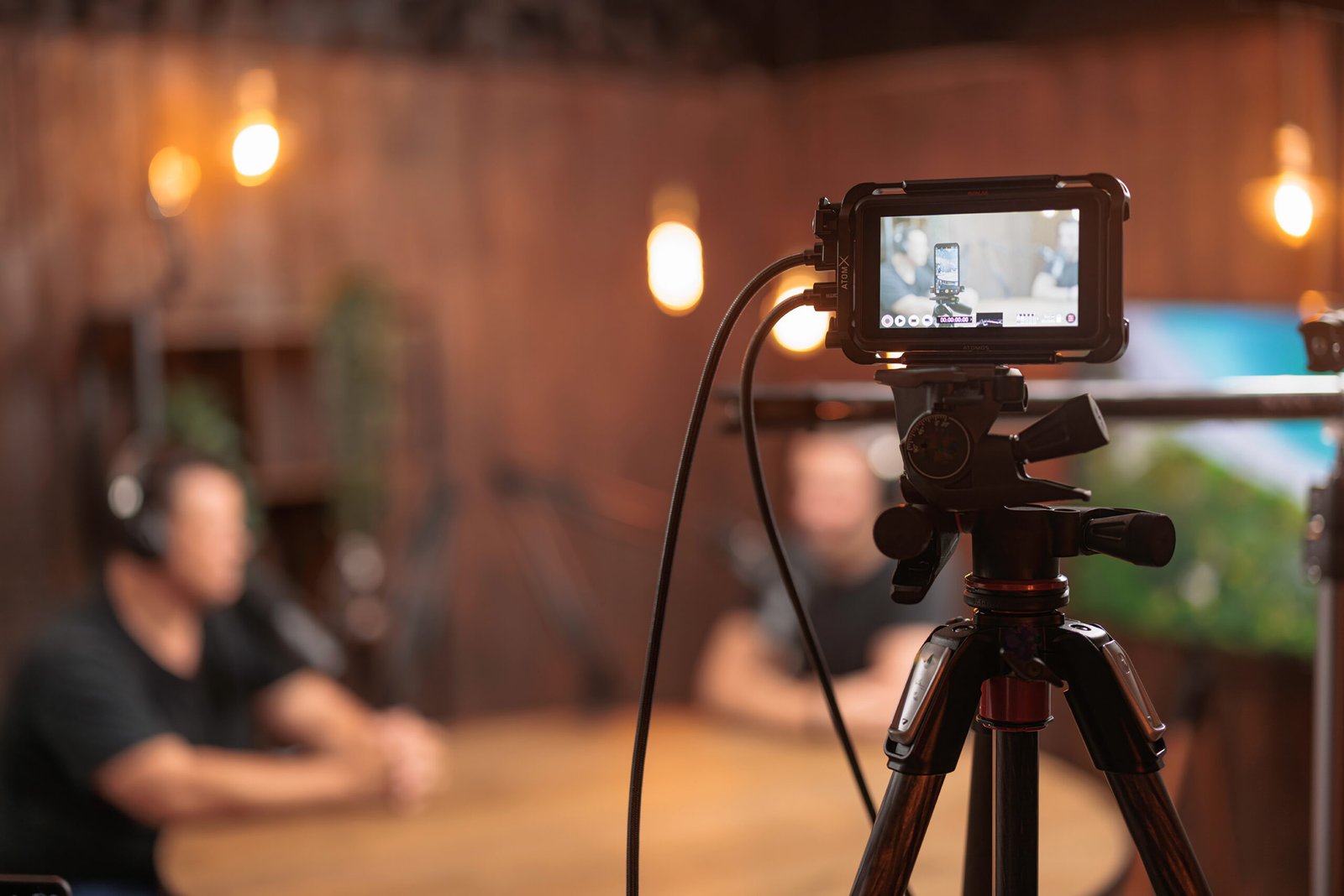 Camera on tripod capturing a podcast recording session with two hosts in a well-lit studio, featuring warm wooden decor and audio equipment.