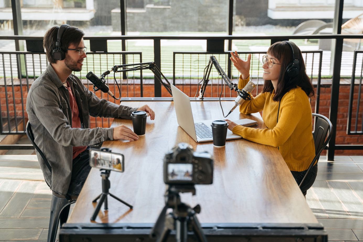 Two podcasters engaged in a discussion at a wooden table, featuring microphones, laptops, and coffee cups, set in a modern studio environment.